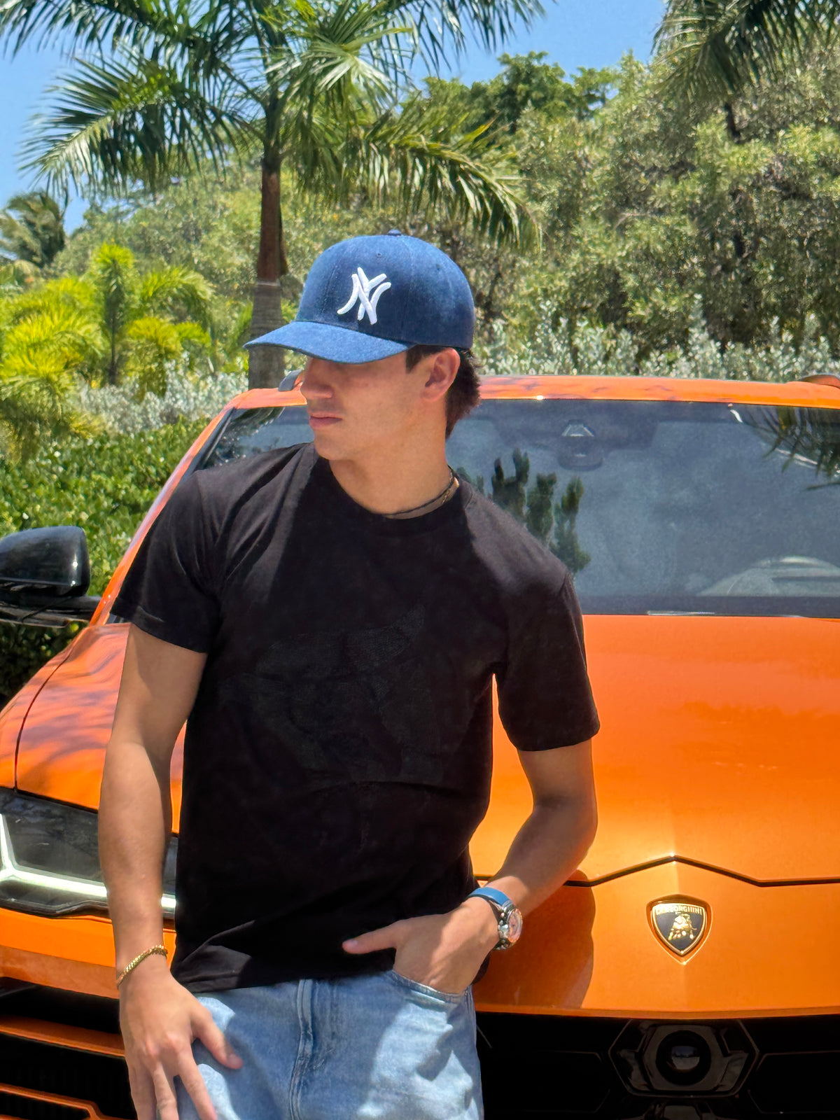 Man wearing the New York denim snapback cap, side profile view, black t-shirt and jeans, orange Lamborghini in background