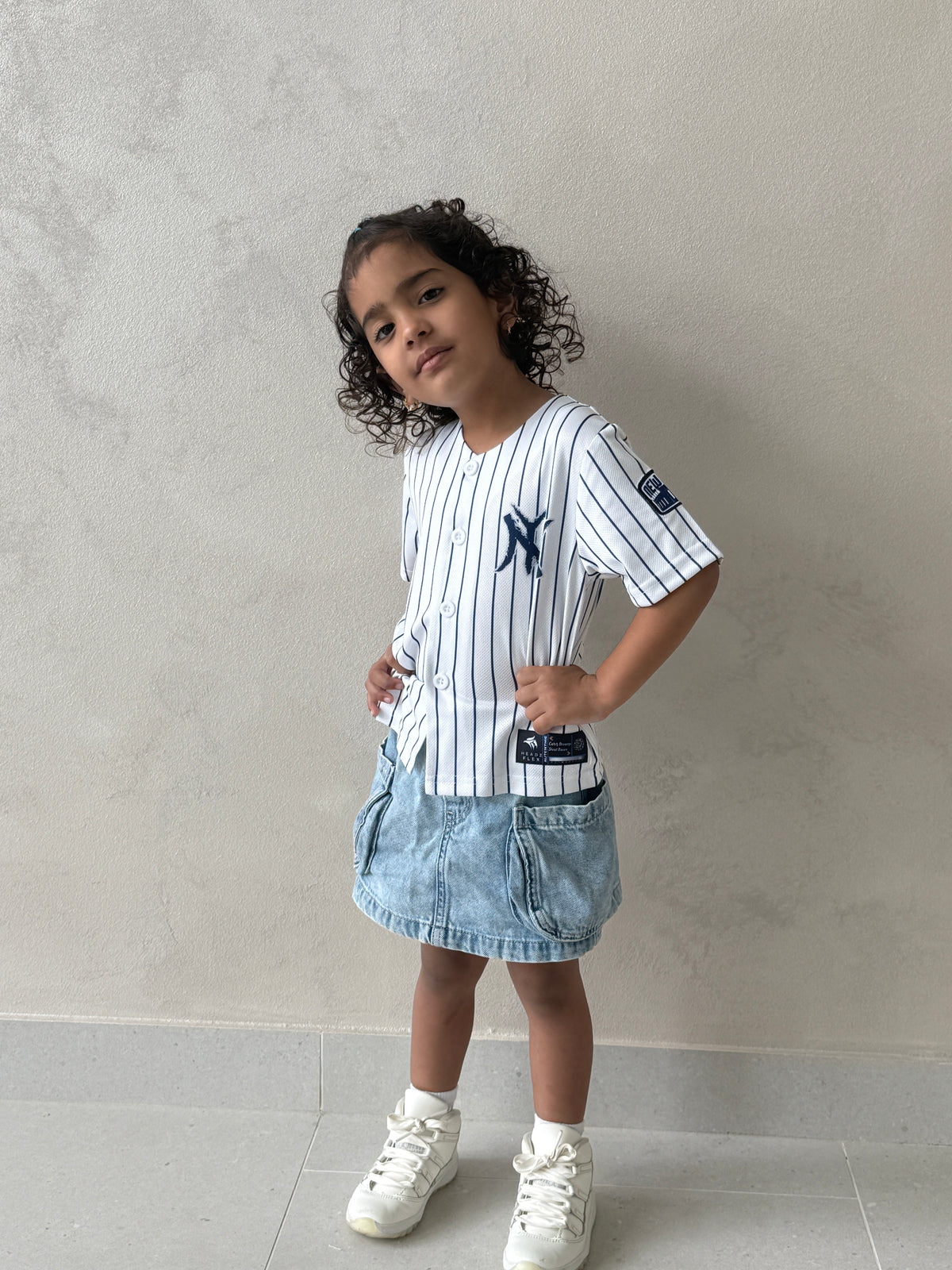 Young girl standing in the white pinstripe New York kids baseball jersey with navy NY logo, denim cargo skirt and white sneakers against a textured gray wall