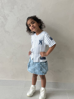 Young girl standing in the white pinstripe New York kids baseball jersey with navy NY logo, denim cargo skirt and white sneakers against a textured gray wall