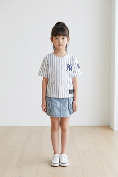 Young girl standing in the white pinstripe New York kids baseball jersey with navy NY logo, denim skirt and white sneakers on a white studio background