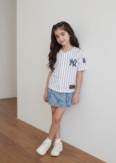 Young girl with long wavy hair wearing the white pinstripe New York kids baseball jersey with navy NY logo, denim skirt and white sneakers against a white wall