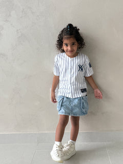 Young girl with curly hair wearing the white pinstripe New York kids baseball jersey with navy NY logo, denim skirt and white sneakers against a wall