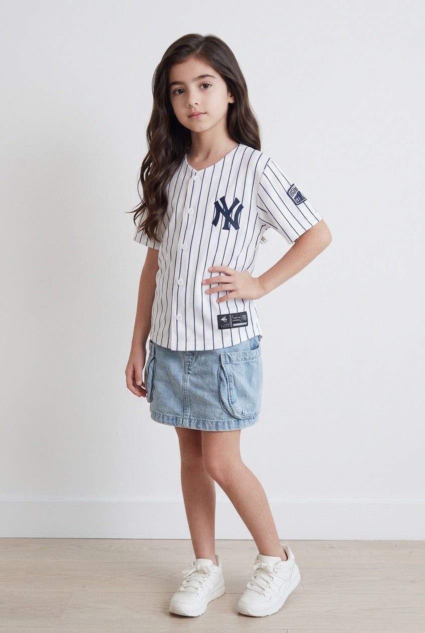 Young girl posing in the white pinstripe New York kids baseball jersey with navy NY logo, denim cargo skirt and white sneakers on a white studio background