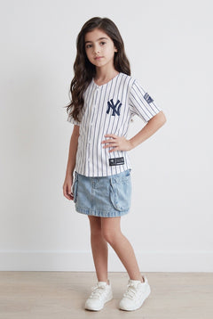 Young girl posing in the white pinstripe New York kids baseball jersey with navy NY logo, denim cargo skirt and white sneakers on a white studio background