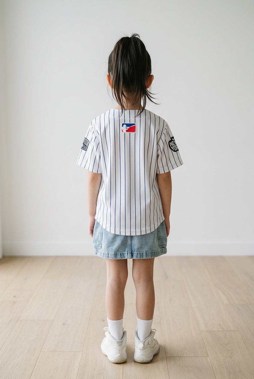 Back view of a young girl wearing the white pinstripe New York kids baseball jersey, denim skirt and white sneakers against a white wall