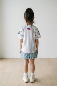 Back view of a young girl wearing the white pinstripe New York kids baseball jersey, denim skirt and white sneakers against a white wall