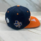 A blue and orange baseball cap with embroidered patches, placed flat on a marble surface.