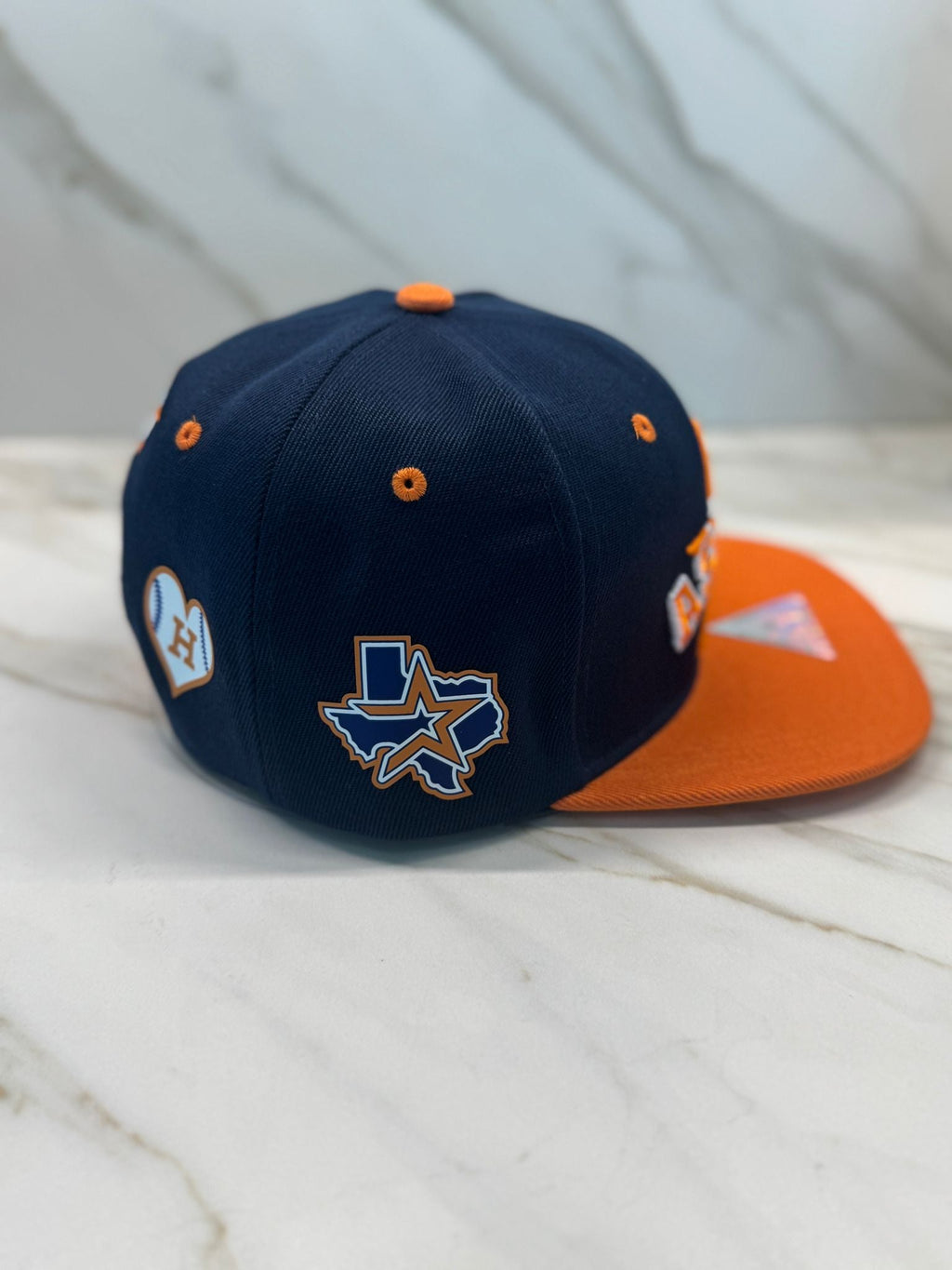 A blue and orange baseball cap with embroidered patches, placed flat on a marble surface.