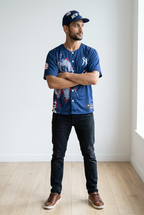 Man wearing a blue baseball jersey and cap in a minimalistic room.