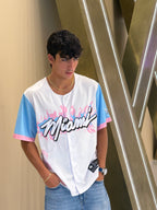 Person wearing a white and blue sports jersey with 'Miami' printed on it, standing against a metallic staircase.