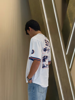 Person wearing a white sports jersey with graphics and text, standing against a modern architectural background.