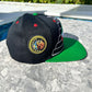 A black and green Headz Flex Cap with embroidered Maya pyramids and a Quetzalcoatl snake on the front, displayed on a flat surface with a background of foliage.