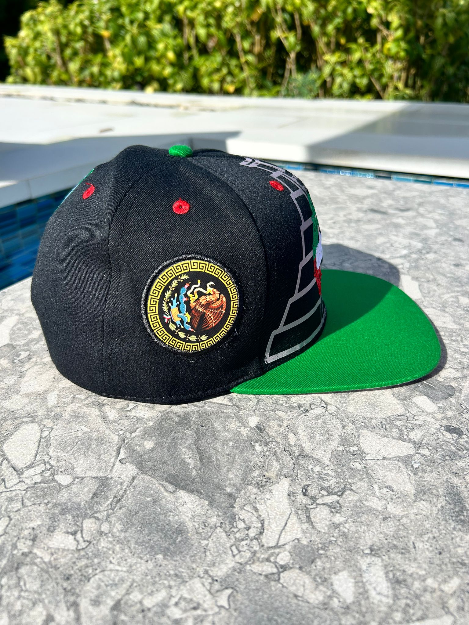A black and green Headz Flex Cap with embroidered Maya pyramids and a Quetzalcoatl snake on the front, displayed on a flat surface with a background of foliage.