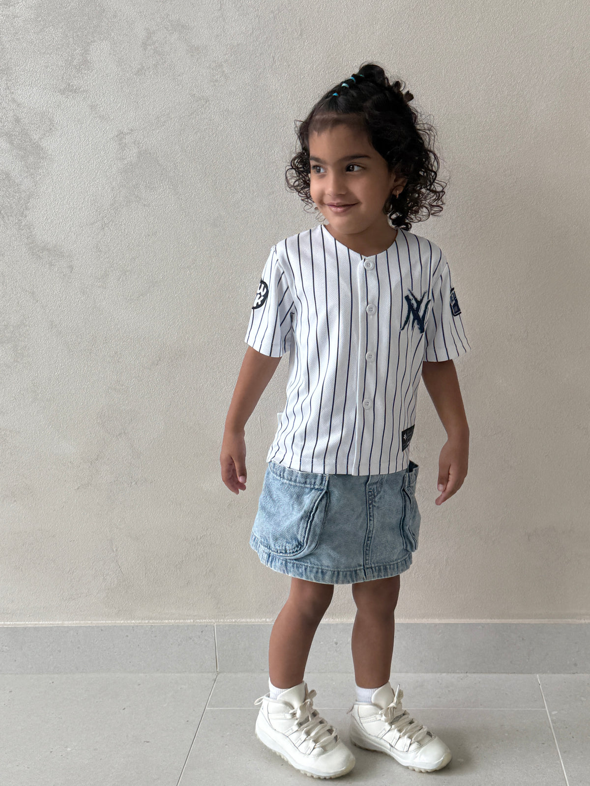 Full-body shot of a young girl wearing the white pinstripe New York kids baseball jersey with navy NY logo, denim skirt and white sneakers against a beige wall