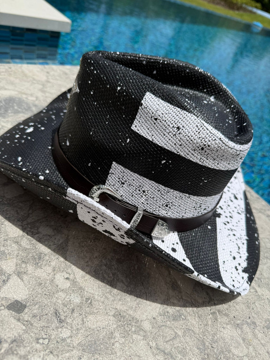 Black and white patterned hat with a black band on a concrete surface near a pool.