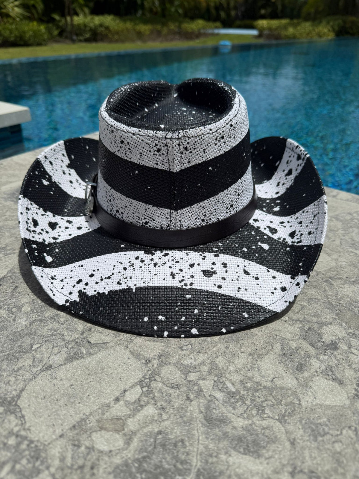 Black and white striped sun hat with splatter pattern on a stone surface near a pool.