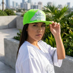 Woman wearing a bright green and white cap with 'Puebla' printed on it, standing outdoors with plants and buildings in the background.