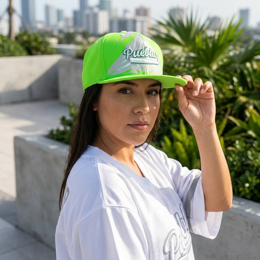 Woman wearing a bright green and white cap with 'Puebla' printed on it, standing outdoors with plants and buildings in the background.