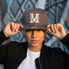 Person wearing a cap with a large 'M' logo against a graffiti-covered wall.
