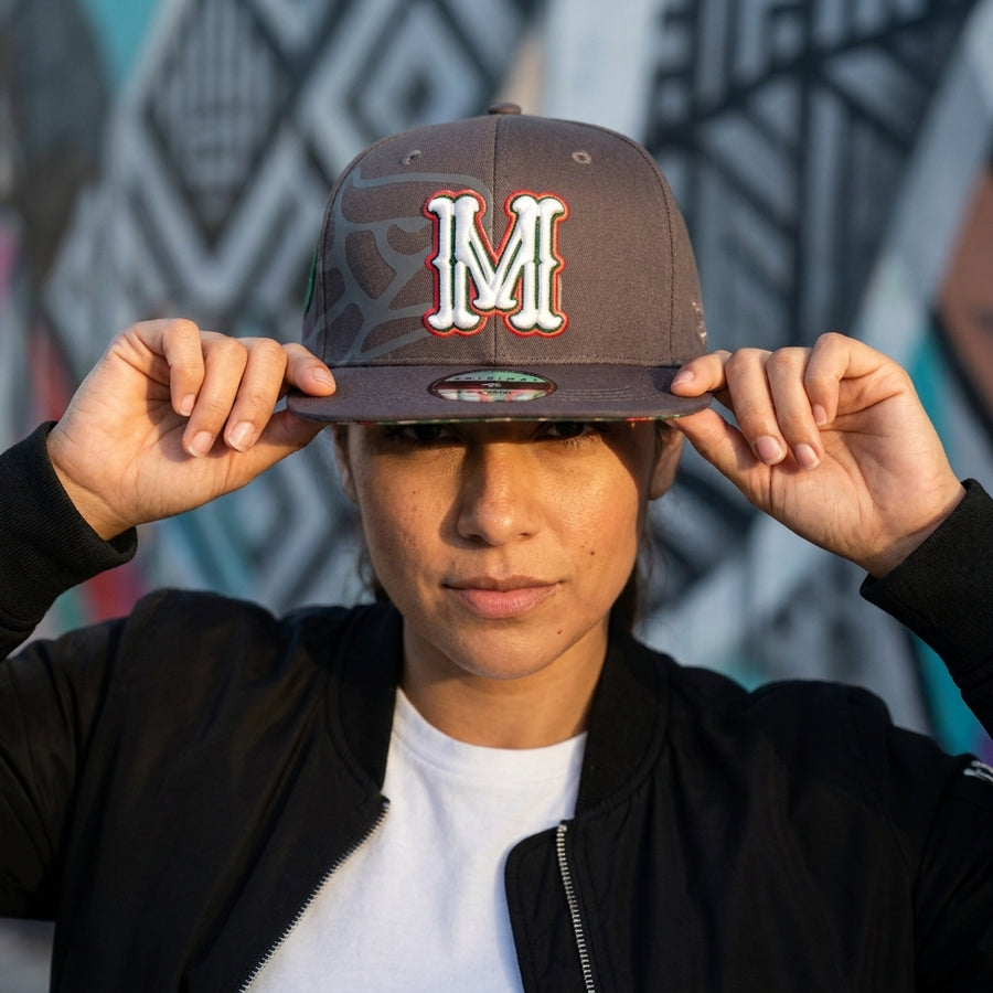 Person wearing a cap with a large 'M' logo against a graffiti-covered wall.