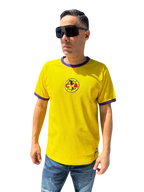 A person wearing a yellow Club America soccer jersey with blue details and a logo on the chest. The jersey has short sleeves, and the person is also wearing sunglasses and blue shorts.