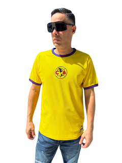 A person wearing a yellow Club America soccer jersey with blue details and a logo on the chest. The jersey has short sleeves, and the person is also wearing sunglasses and blue shorts.