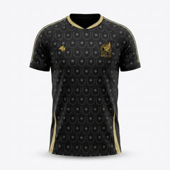 A black short-sleeve soccer jersey with a gold and gray repeating scale pattern, featuring a gold chest crest and a small gold logo on the right chest, displayed on a light gray background.